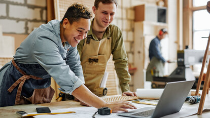 Two craftsmen check plans on a laptop.