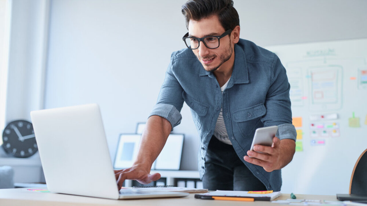 Mann hält Handy in der Hand und arbeitet am Laptop