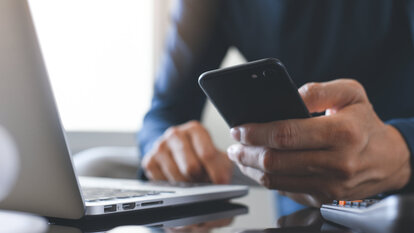 A man holds a smartphone in his hand and sits at his laptop