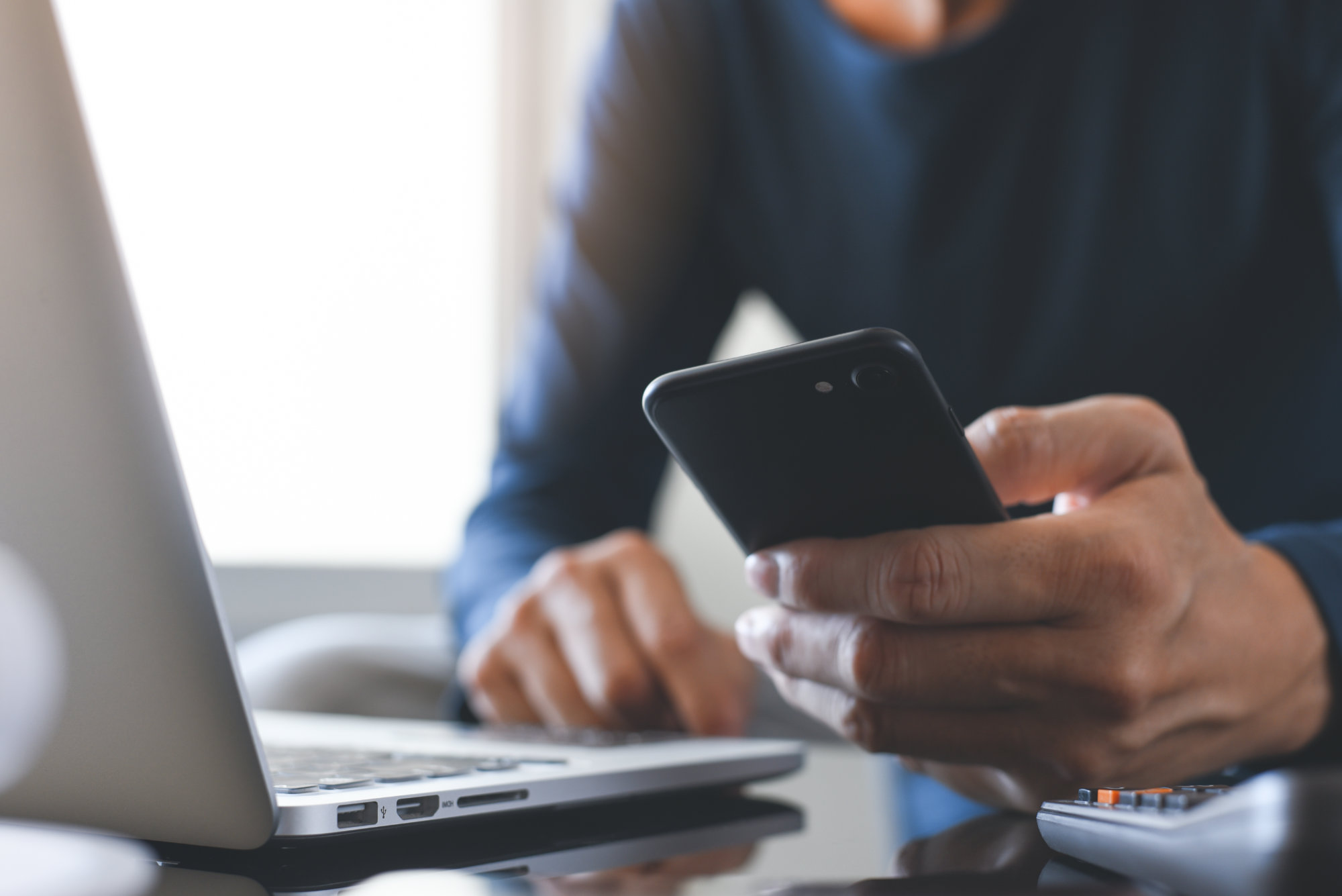 Mann hält ein Smartphone in der Hand und sitzt vorm Laptop.